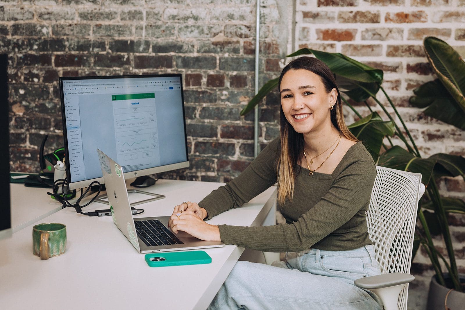 Meara smiles at the camera while preparing a Google Looker Studio report on her computer in the office.