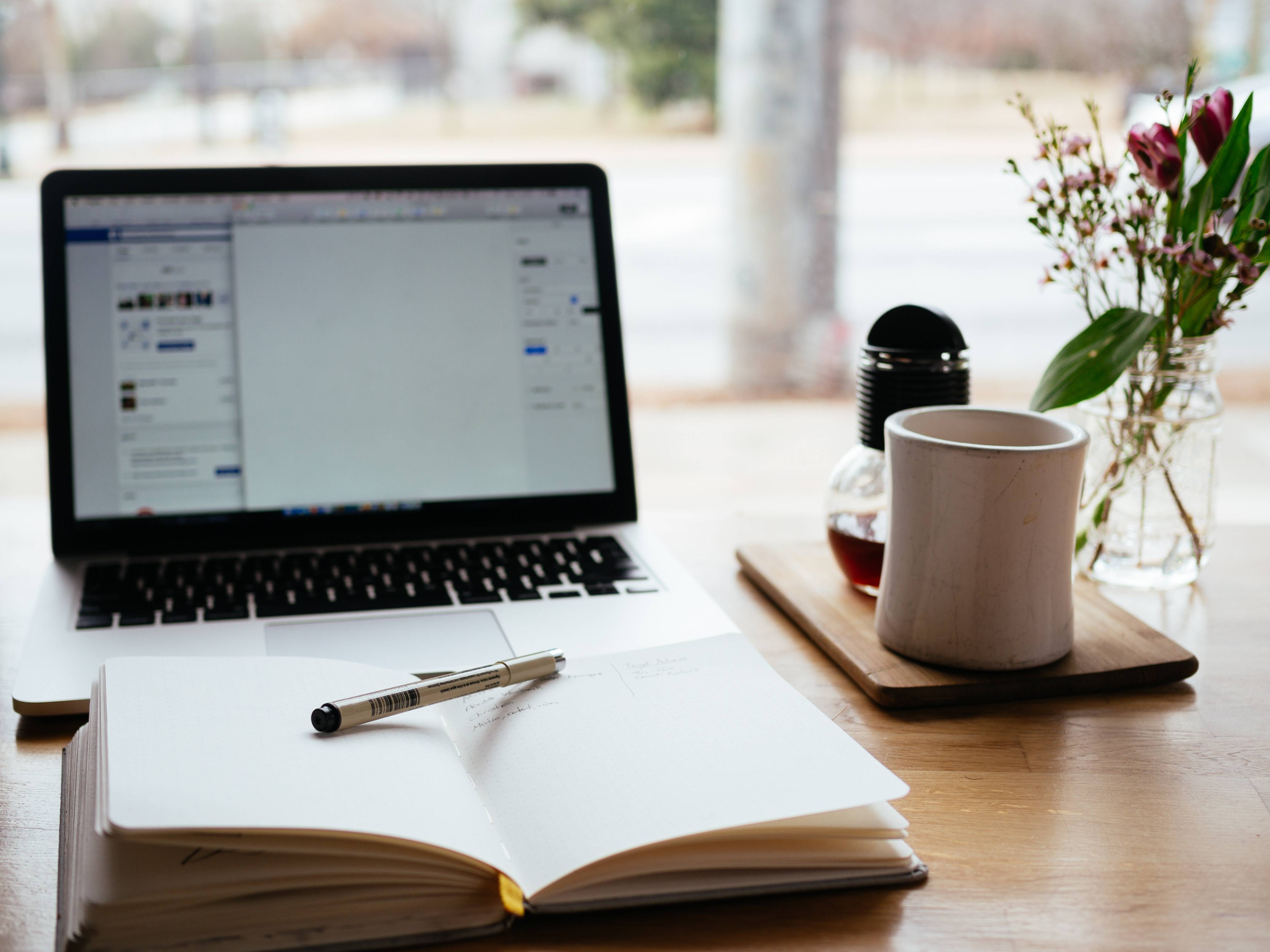 A typical scene for someone sitting down to write a blog post: an open notebook, laptop, and cup of coffee on a desk. Knowing your purpose before you write is one way to improve your blogging.