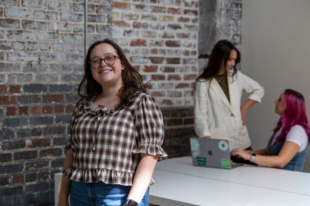 Abby stands smiling in the office. Behind her, Meara and Madison are in conversation.