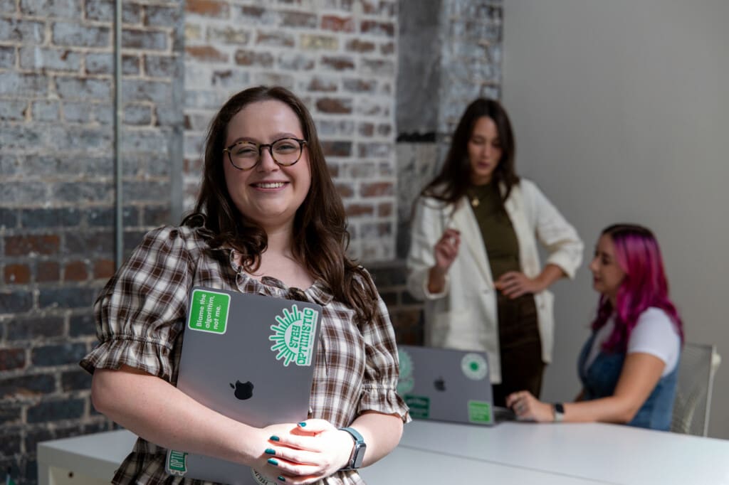 Abby stands, smiling and holding her laptop, in the office. Behind her, Meara and Madison have a conversation.