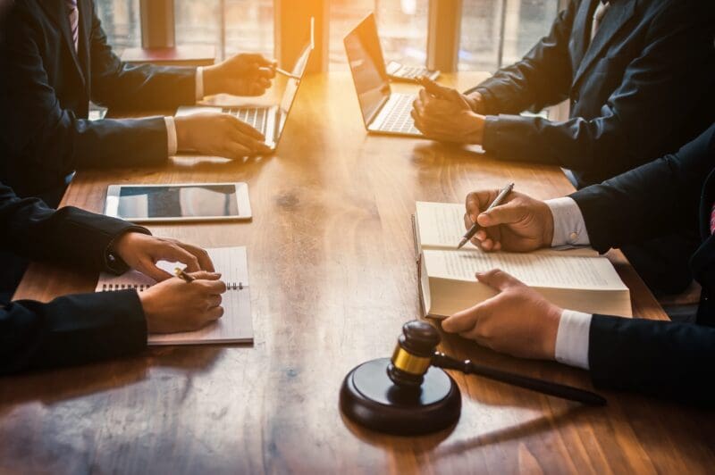 People sit at a wooden table together and discuss over books, notebooks, and laptops. There is a gavel in the foreground.