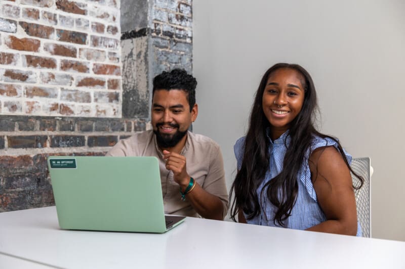 Esteban and Kimberly sit next to each other in the office. Esteban works on a laptop, and Kim smiles at the camera.