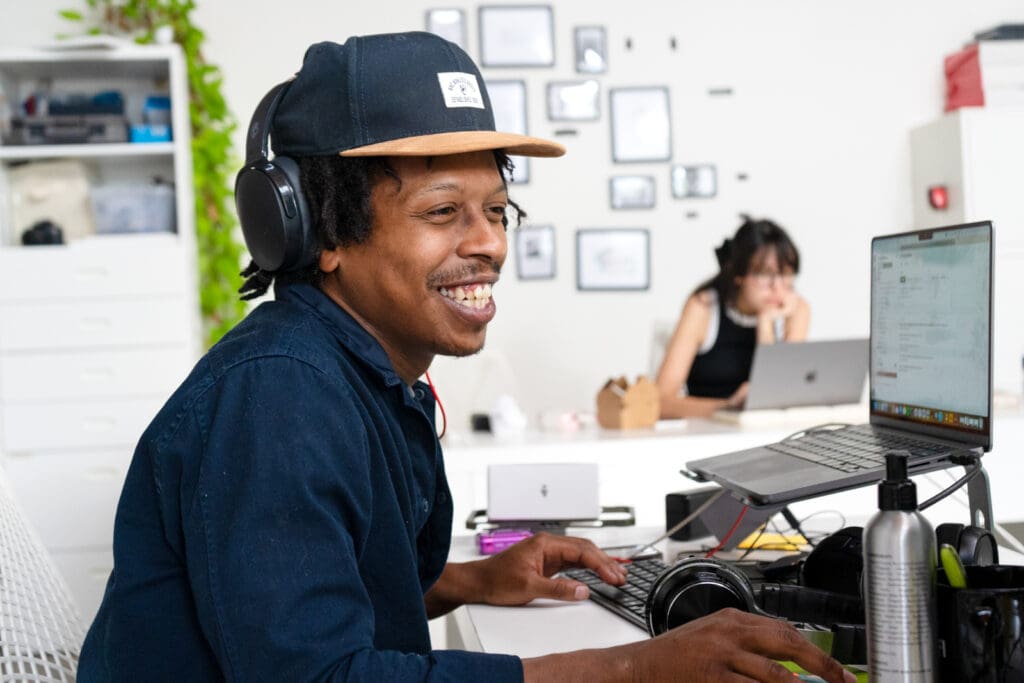 Daniel smiles while working in the Online Optimism office, with Jordan deep in thought at a desk behind him.