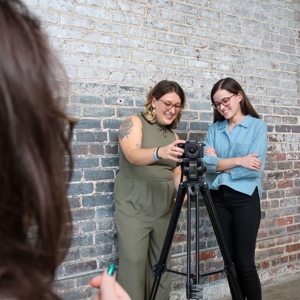 Erin and Claire stand behind a camera during a video shoot. Erin adjust the camera.