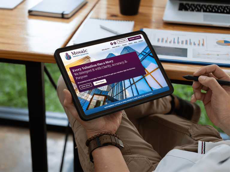 Professional man sits at desk holding tablet displaying Mosaic Property Valuations' website homepage