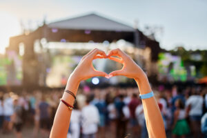 A fan wearing friendship bracelets makes a heart with their hands in the air at a live concert.