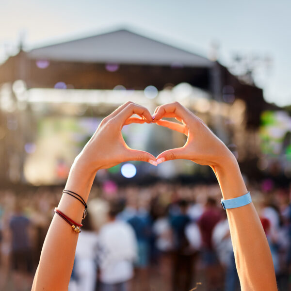 A fan wearing friendship bracelets makes a heart with their hands in the air at a live concert.