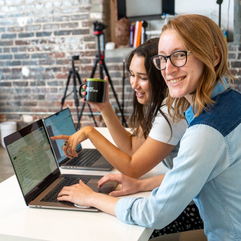 Lauren and Meara work on laptops side by side. Lauren looks up at the camera and smiles as Meara points to something on the screen.