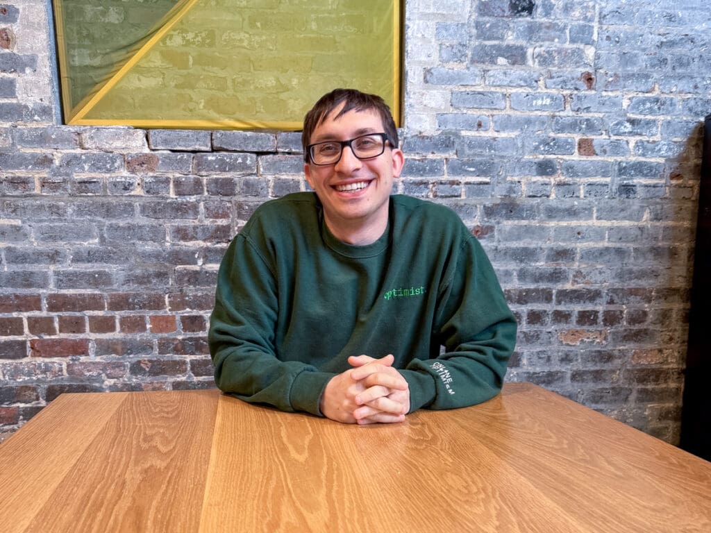 Flynn sits at the table in the Online Optimism office and smiles at the camera.