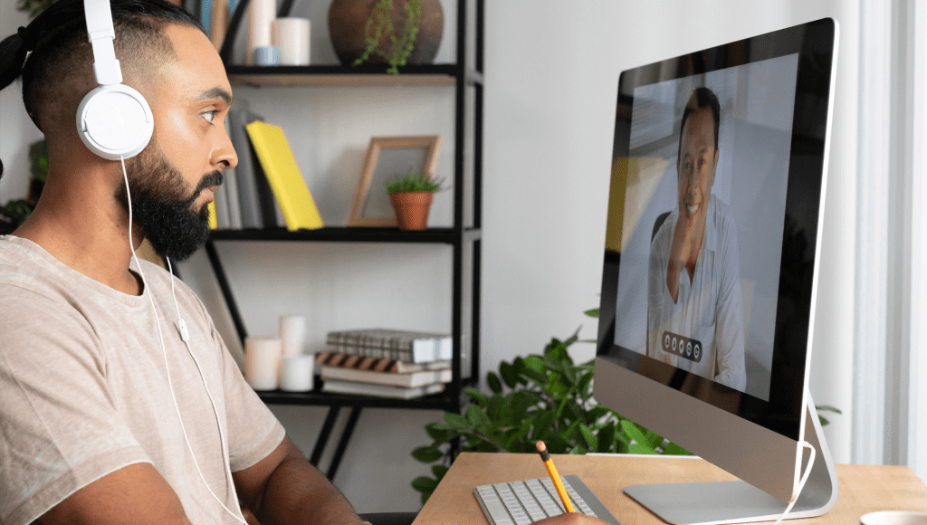 A man listens to a video call in his home office with headphones on and takes notes.