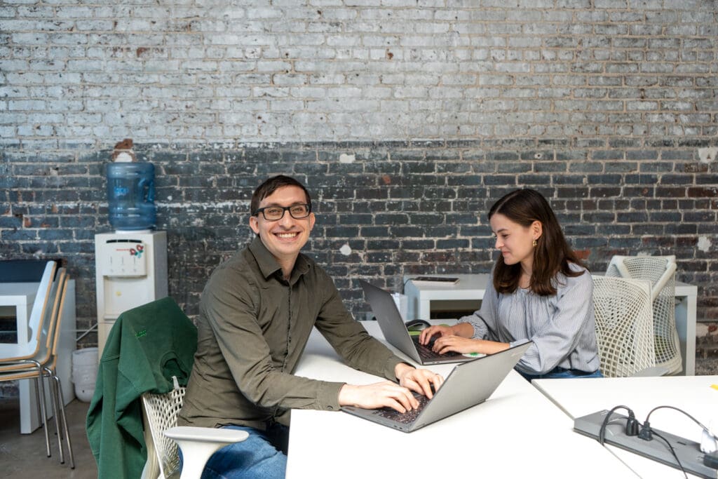 Flynn and Claire work on their laptops in the Online Optimism office. Flynn looks up at the camera with a smile.