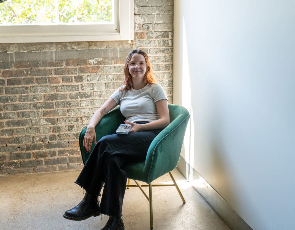 Bri looks up from her phone, sitting in a green chair at the Online Optimism office.