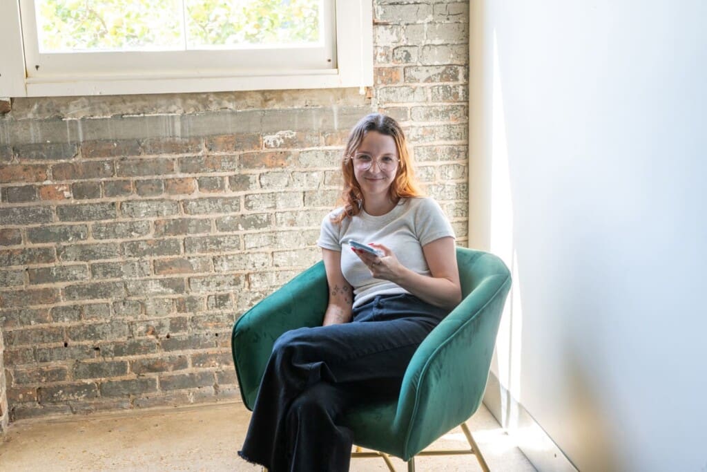 Bri sits in a chair in the office. She looks up from her phone to smile.