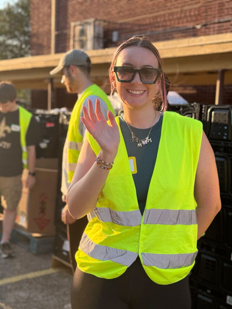 Madison waves at the camera while volunteering for Culture Aid NOLA, wearing a yellow safety vest like other volunteers in the background.