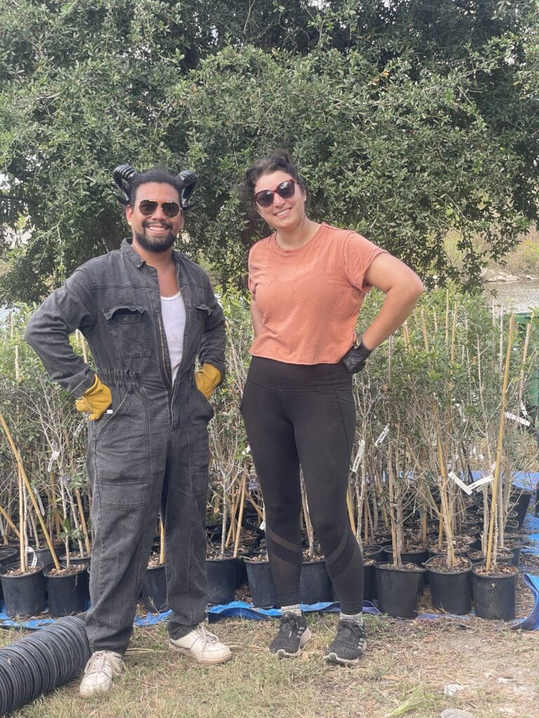 Sara and Esteban stand together outside next to rows of new baby trees in pots, smiling for the camera.