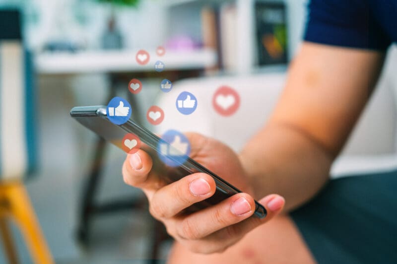 A person holding a phone, with Facebook-style hearts and thumbs-up emojis floating off it.
