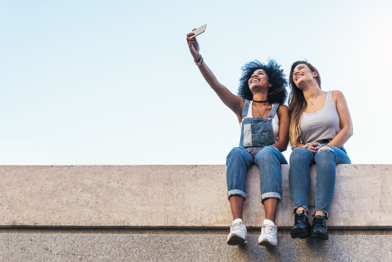 Two people sit side-by-side on a concrete wall and take a selfie together.