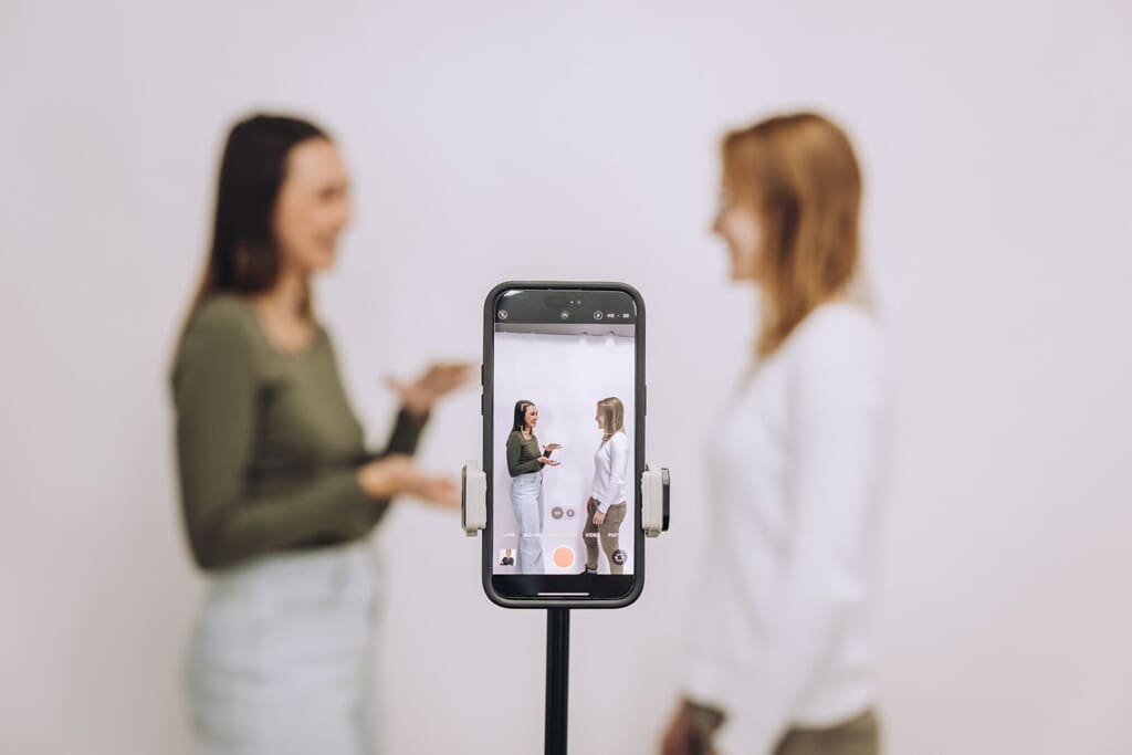 In the foreground, a smartphone takes a video of Meara and Lauren, who are out of focus against a white background