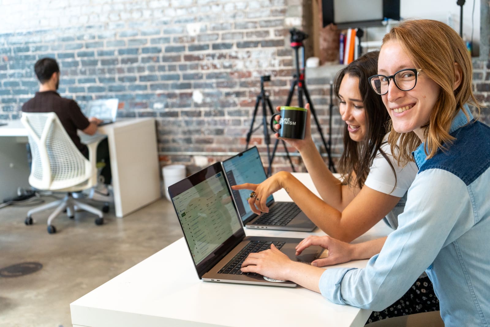 Lauren and Meara work on laptops side by side. Lauren looks up at the camera and smiles as Meara points to something on the screen.