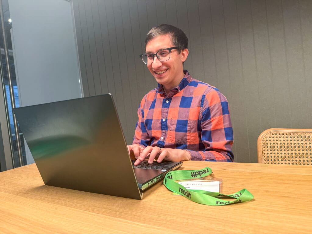 Flynn smiles while working on a laptop at an office table. An official Reddit moderator lanyard is on the table next to him.