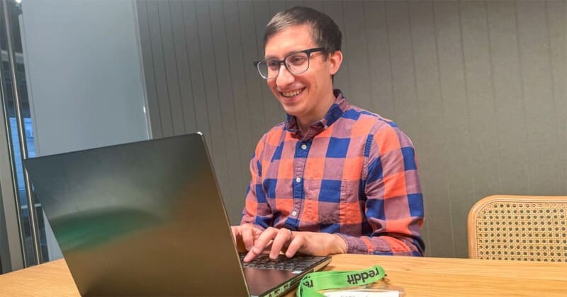 Flynn smiles while working on a laptop at an office table. An official Reddit moderator lanyard is on the table next to him.