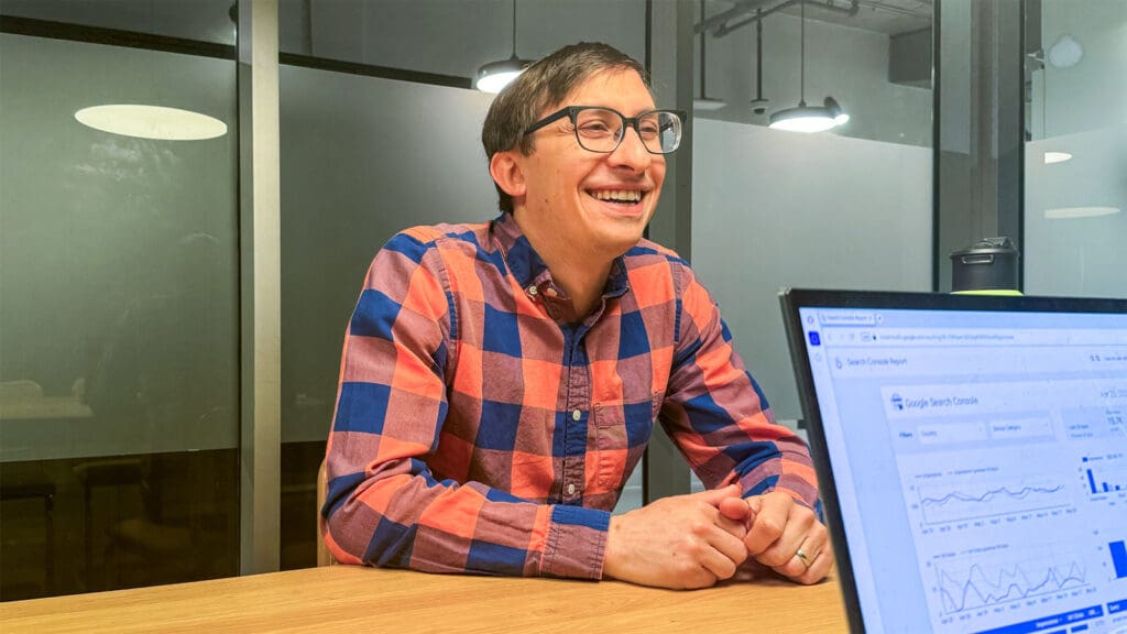 Flynn sits at an office table, smiling as he talks to a person off camera who uses their laptop to look at a Google Search Console screen