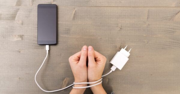 A photo of a person's hands being wrapped by a smartphone cord, as if shackled. Learn how to overcome social media addiction with our guide.