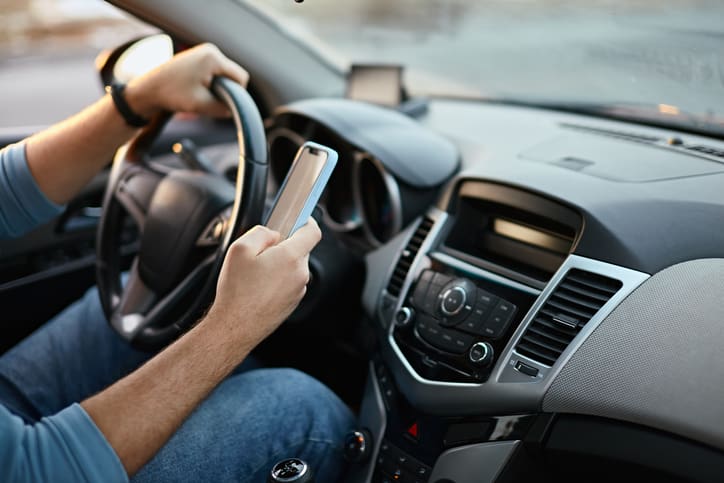 A photo of a driver who is looking at their phone while holding the steering wheel