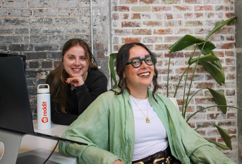 Natasha and Hannah look up from work on Reddit and smile. A Reddit water bottle is on their desk.