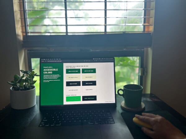 Photo of workspace with laptop in front of window. The laptop screen displays a page of a brand guide with a chart of accessible colors.