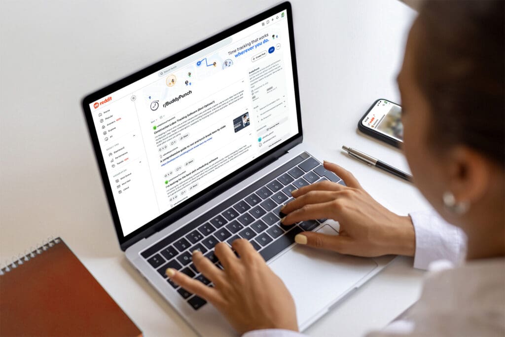 Person uses a laptop to view Buddy Punch's subreddit. A phone with the Reddit app open and a red notebook are also on the desk.