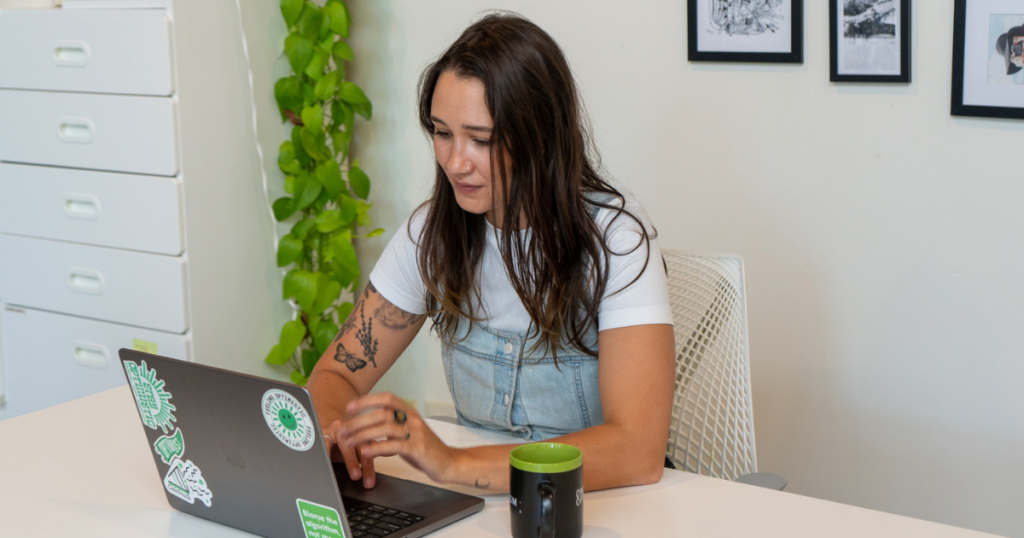 Meara McNitt, VP of People at Online Optimism, working on a laptop at a white desk with a coffee mug, houseplant, and gallery wall of framed artwork in the background.