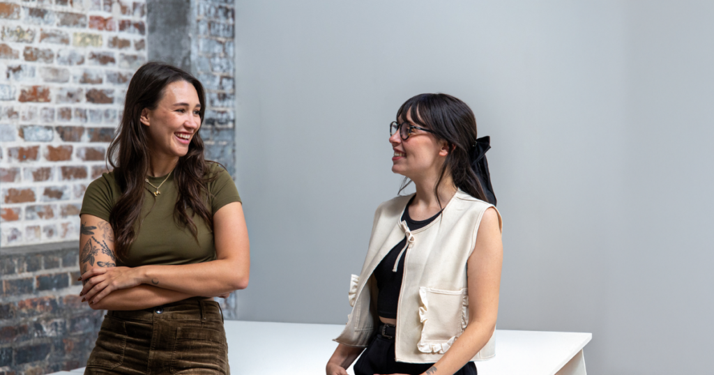 Two women smiling and chatting in a bright room with a brick wall background. One wears a green top and has a tattoo, the other wears glasses and a white vest.