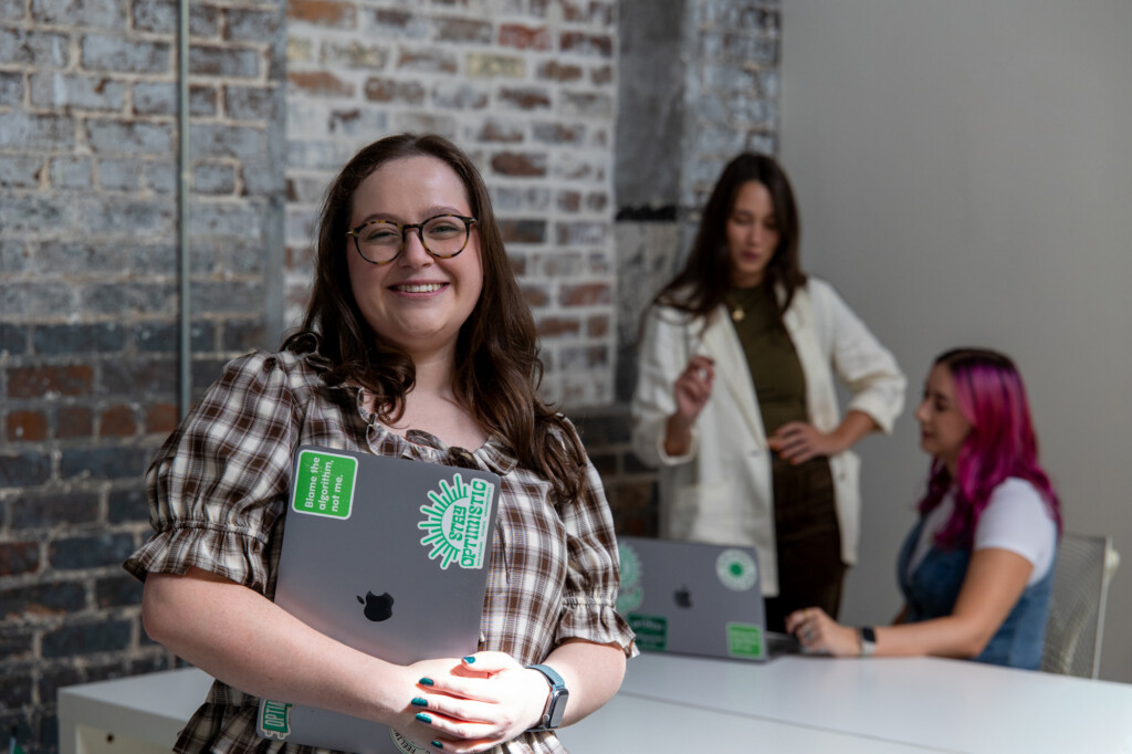 Abby, our Social Media Strategist, poses with her laptop while Madison and Meara do social media work in the background.