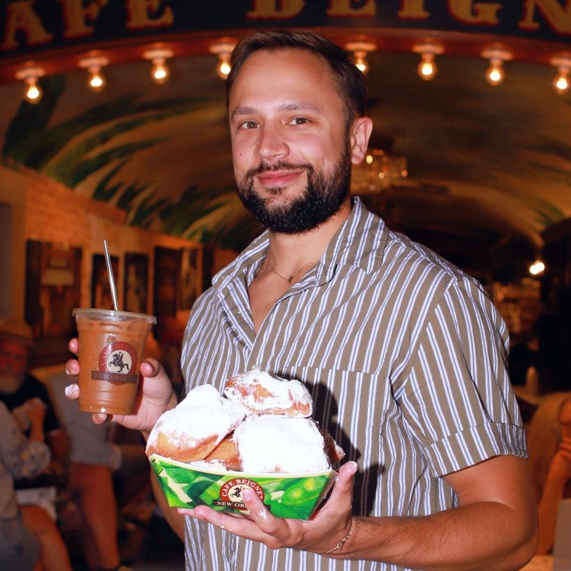 New Orleans influencer Big Eatsy (Mattlan Ladner) smiles at the camera as he holds a towering paper plate of beignets and an iced coffee from Cafe Beignet.