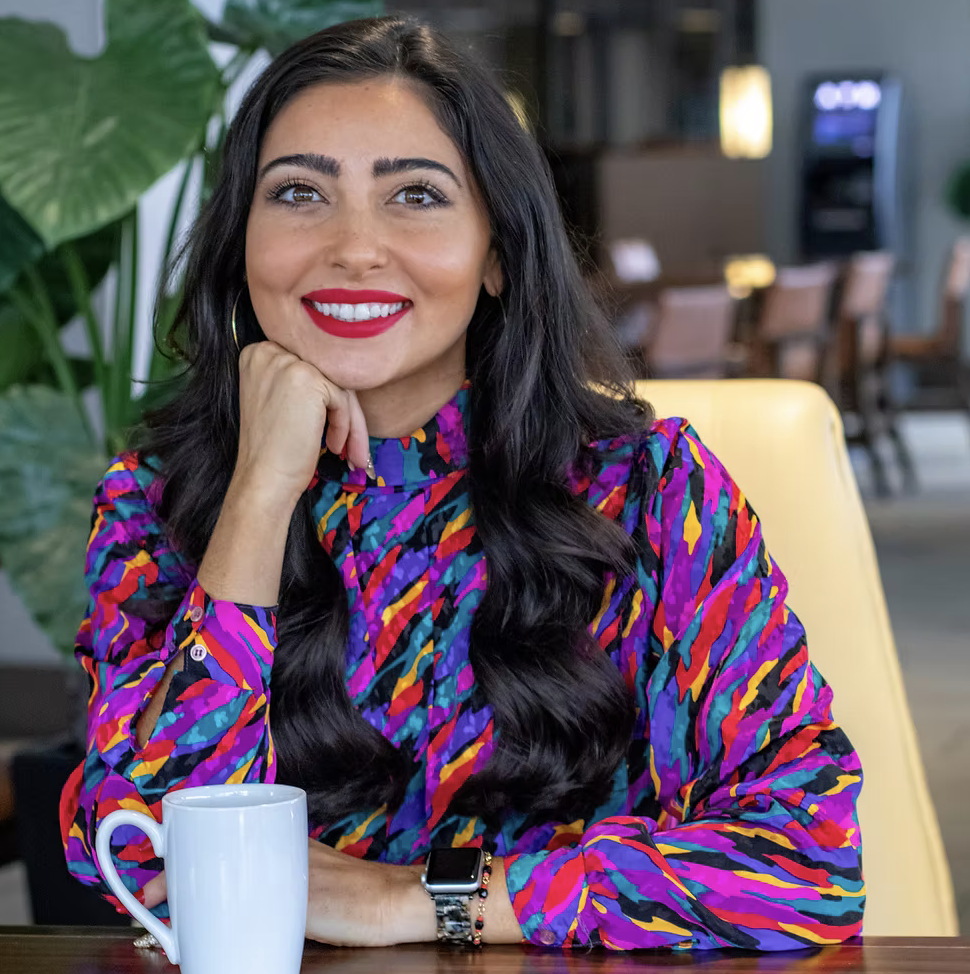 New Orleans influencer Eaten Path NOLA (Nicole Caridad Ralston) smiles for the camera as she rests her chin on her hand, wearing a colorful blouse.