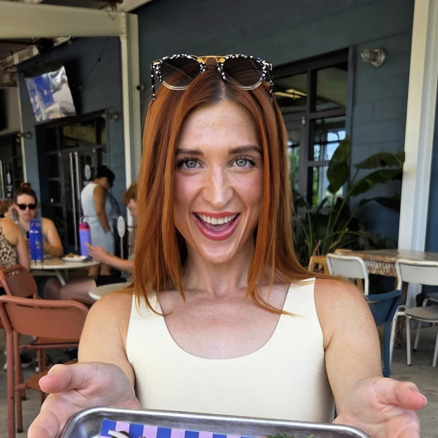 New Orleans influencer Five Oh Pour (Kayla McCormick) holds a tray of food up to the camera outdoors on a restaurant patio with a big smile.