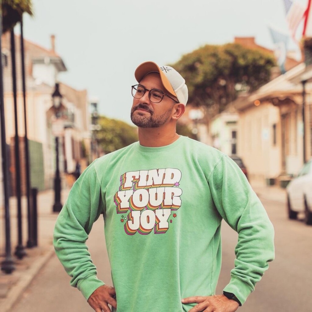 New Orleans influencer Gregisms (Greg Kata) stands outside wearing a green sweatshirt that says "Find Your Joy" in layered rainbow block letters.
