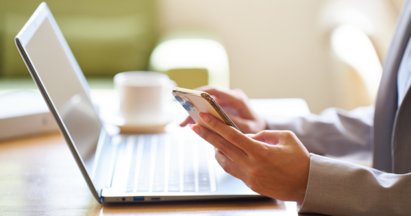 A marketer uses their smartphone to share content on social media with their laptop open in the background where content is being distributed across other channels